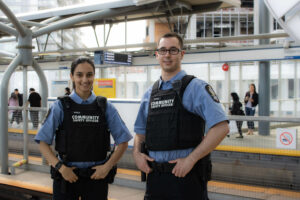 Two Community Safety Officers, a young woman and a young man, smile at the camera while standing at Sapperton SkyTrain Station.