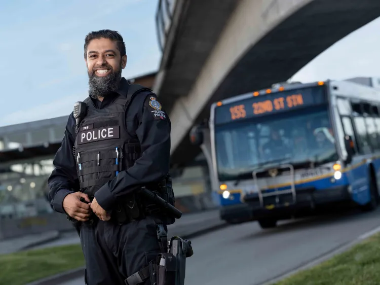 Transit Police officer Constable Shiraaz Hanif smiles at the camera with a bus and SkyTrain station in the background