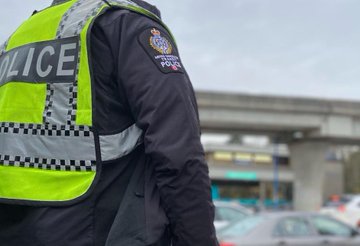 A Transit Police officer is standing facing traffic with a SkyTrain station in the background